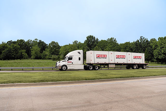 A white PODS tractor trailer driving on a highway. The truck is hauling PODS storage containers